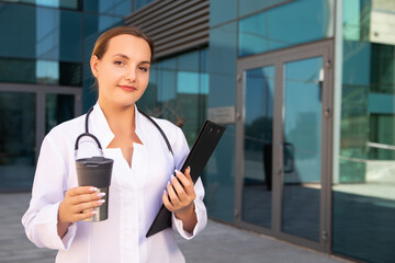 Horizontal photo of cute female therapist with stethoscope around her neck. Doctor looking the camera, holding the test folder and her coffee cup. Copy space. Medicine, people and healthcare concept.