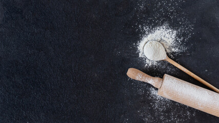 Wheat flour in a wooden spoon with rolling pin over black surface top view.