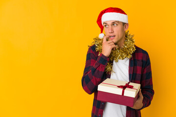 Young caucasian man with christmas hat holding a present isolated on yellow background