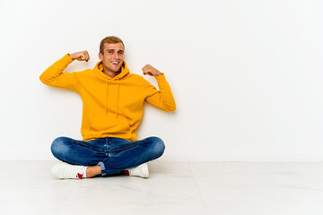 Young caucasian man sitting on the floor showing strength gesture with arms, symbol of power