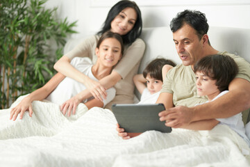 Days at home. Father using tablet pc, watching cartoons, playing games with two little sons. Happy latin family with children staying in bed together in the morning