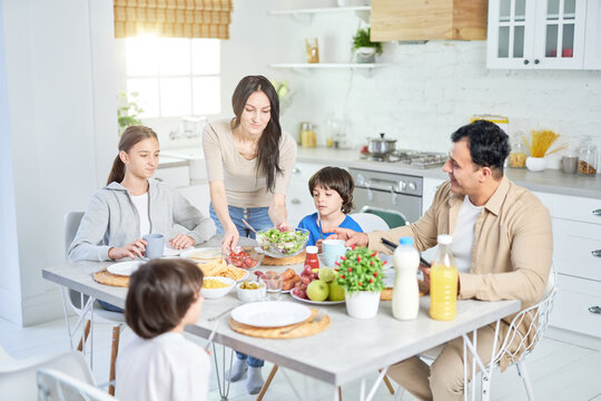Share food. Caring hispanic woman serving salad for her husband and children, standing in the kitchen. Latin family having dinner together at home