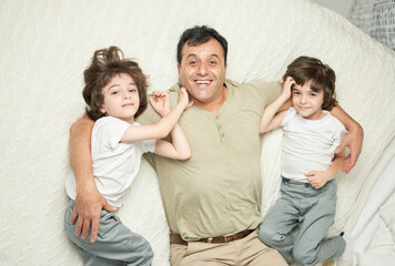 Two cute little latin boys, children and their father smiling at camera while lying together on a bed at home