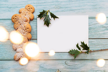 gingerbread cookies, gingerman, arranged on a blue table with holly, traditional Christmas plant, Christmas lights bokeh, and a blank white card with copyspace, lets you create a tasty Christmas card - 396662935