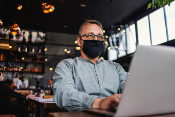 Middle aged serious freelancer with eyeglasses and protective mask on sitting in a cafe and using laptop during corona outbreak.