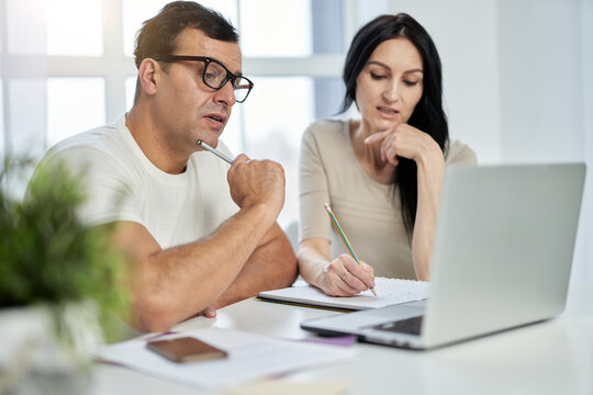 Learning At Your Own Convenience. Middle Aged Latin Couple, Man And Woman Making Notes During Online Training Course While Studying Remotely, Using Laptop At Home