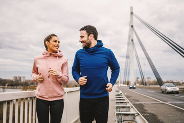 Smiling happy sporty couple jogging on the bridge at cloudy autumn day.