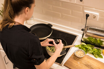 Rear view of a young beautiful girl cooking and preparing vegetarian food in her kitchen alone. Swedish woman grinding pepper into the pot while she's preparing a healthy recipe for the family