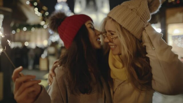 Charming girls lesbians couple meeiting and hugging at festive street fair on New Year's Eve. LGBT community representatives pleasantly having time, enjoing atmosphere of holiday and burning sparklers