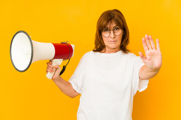 Middle age caucasian woman holding a megaphone standing with outstretched hand showing stop sign, preventing you.