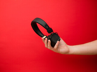 A young woman holds a pair of black wireless headphones in her hand. Hand of a girl with headphones on a red background. Musical concept.