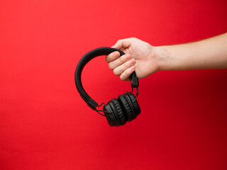 A young woman holds a pair of black wireless headphones in her hand. Hand of a girl with headphones on a red background. Musical concept.