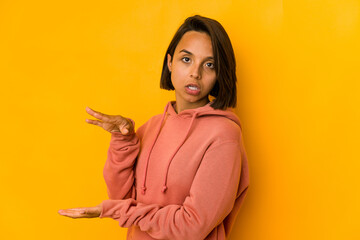 Young hispanic woman isolated on yellow shocked and amazed holding a copy space between hands.