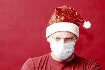 Grumpy man in Santa hat and medical protective mask on a red background.
