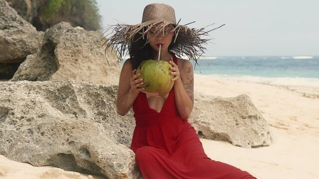 Beauty, baunty, travel, fashion concept. Woman quench her thirst with coco nut. Vacation on exotic island. Beautiful pretty asian woman in hat and red dress drinking coconut on tropical paradise beach