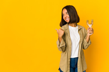 Young hispanic woman holding a slingshot points with thumb finger away, laughing and carefree.