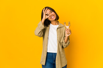 Young hispanic woman holding a slingshot excited keeping ok gesture on eye.