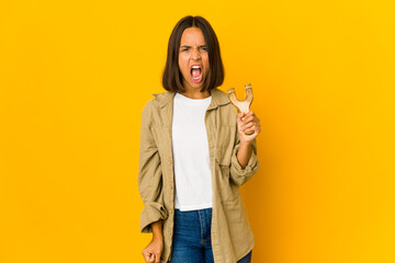 Young hispanic woman holding a slingshot screaming very angry and aggressive.