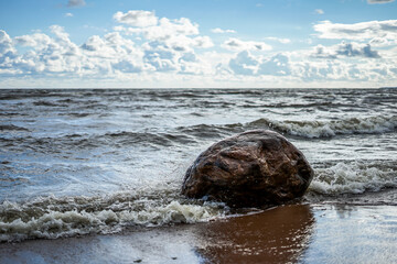 The wave beats against a large stone on the seashore, a clear day, light clouds