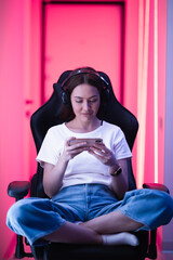 Cybersport gamer playing mobile game on the smart phone sitting on a gaming chair in neon color room. - 396675566