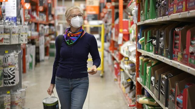 Slow motion of mature woman wearing face mask holding paint and walking down aisle in hardware store. Concept of coronavirus shopping experience.