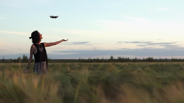 Pretty woman controls drone fly to the sky in summertime sunset in the field. Girl set copter from hand standing in tall grass. Autimn wheat field and a person. technology, gadget, internet concept.