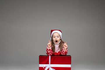 Studio shot of pretty little girl in Santa hat and winter sweater announcing information with fingers up leaning on Christmas present. Isoolate on grey background.
