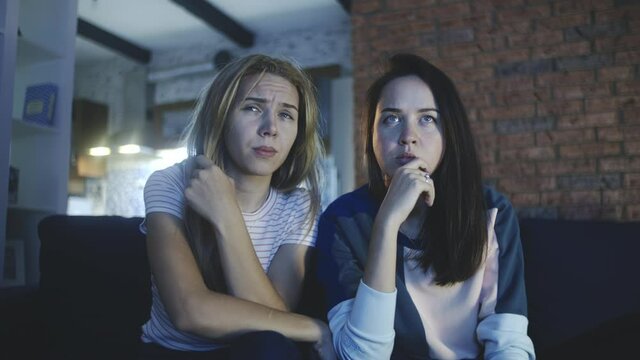 Female friends watch TV together in evening. Two girls watching scary movie or bad and unpleasant news at home. Brunette and blonde sitting on sofa in silence and afraid. Concept of cinema, home party