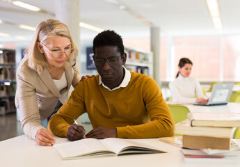 Female teacher working with african-american man in university library. High quality photo