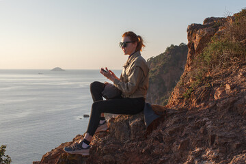 young smiling traveler woman sitting on rocks on sea coast with smartphone