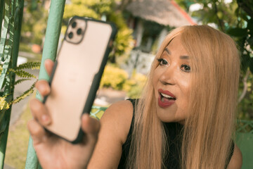 A middle aged single asian woman with dyed straight blonde hair poses for a selfie. Feeling young, sexy and confident. Outdoor setting.