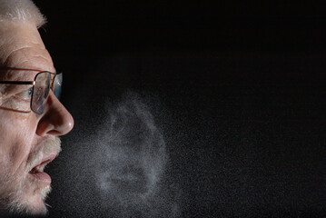 A man sprays aerosols into the air while speaking. The background is black.