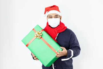 A portrait of a young asia man wearing a face mask and a Santa Claus hat standing in front of a white background and holding a Christmas gift box