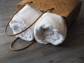 A handmade brown bag made from natural materials have two white towels that are rolled up and placed on a wooden floor by a sunny window.
