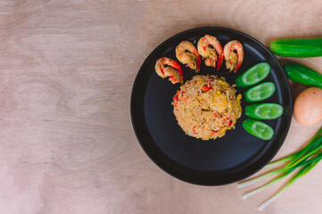 Shrimp fried rice in a black plate with spring onion, egg and cucumber all on the wooden table. top view.
