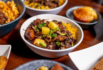 Grilled Pineapple Chicken with Green onion garnish, rice and fried plantains in a white bowl and other food blurred in the background