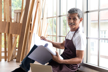 Senior asian elderly old man carpenter study and check blueprint plan for building diy furniture in workshop