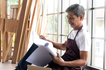 Senior asian elderly old man carpenter study and check blueprint plan for building diy furniture in workshop