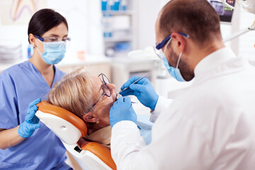 Stomatolog and nurse treats teeth of senior woman using drill. Elderly patient during medical examination with dentist in dental office with orange equipment.