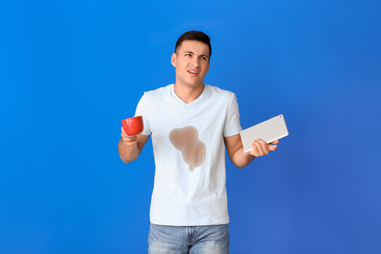 Stressed young man with coffee stains on his t-shirt holding tablet on color background - Powered by Adobe