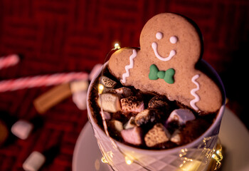Delicious cup of hot chocolate inside a white cup with a gingerbread man cookie dipped in it with chocolates on top sprinkled with cinnamon on a red background and Christmas lights