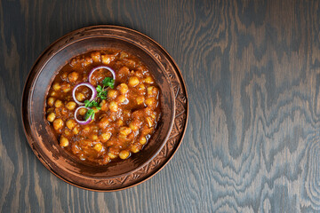 Top view of chole masala or chana indian food made of cooked chickpeas, tomatoes and cumin decorated with onion rings and parsley served in bowl on dark brown wooden background. Image with copy space