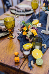 Detail of the wooden table with bouquet, ocher and trasparent glasses, candles, fruits and grey fabric