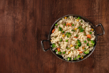 Stir-fried vegetable rice with broccoli, green peas and carrots, overhead shot on a dark rusitic wooden table with a place for text