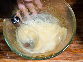 cooking sweet sponge cake at home - above view of whipping of raw egg in glass bowl by electric blender on old wooden table at home kitchen (mixer whisk blurred in motion)