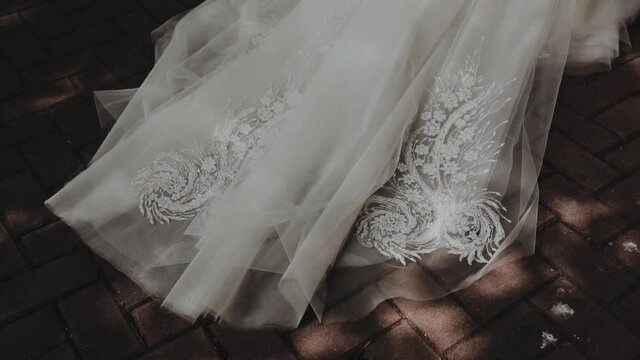 The bride with a long white veil of fatin and expensive lace are going forward and pulls the veil on the tiles in the yard on a sunny day and against the background of shadows from the leaves of trees