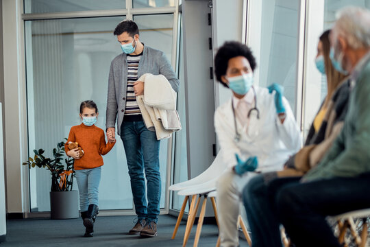 Father And Daughter Wearing Protective Face Masks While Holding Hands And Walking Through Hospital Hallway.