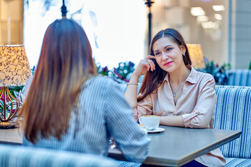 Women are talking over a cup of coffee at a table in a cafe and smiling.