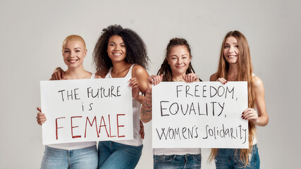 Four happy diverse women smiling at camera while holding, standing with banners in their hands isolated over grey background. Feminism and equality concept