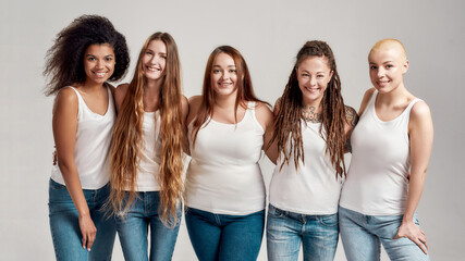 Group of five beautiful diverse young women wearing white shirt and denim jeans smiling at camera while posing together isolated over grey background